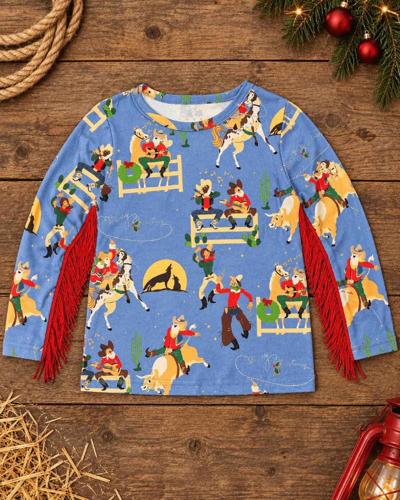 A long-sleeve children's top in light blue with a festive cowboy and horse pattern, featuring red fringe trim along the sleeves, laid flat on a rustic wooden surface with holiday decorations.