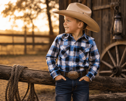 Child wearing a cowboy hat and plaid shirt standing by a wooden fence with a sunset in the background.