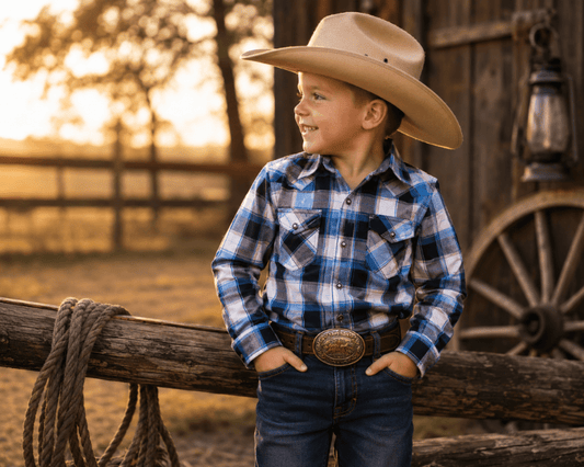Child wearing a cowboy hat and plaid shirt standing by a wooden fence with a sunset in the background.