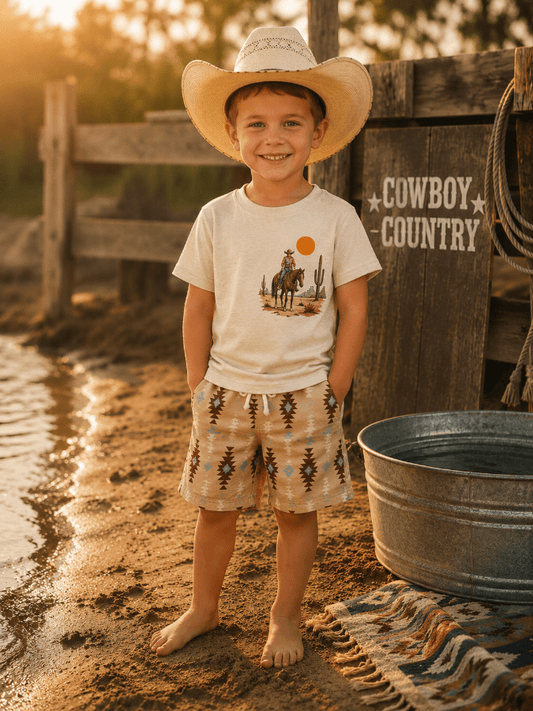 Young boy in a cowboy hat and shirt standing near a wooden sign with 'Cowboy Country' on a dirt path.