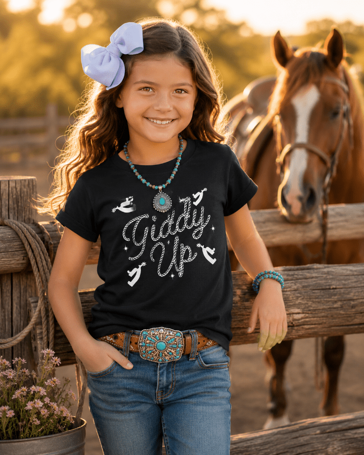 Young girl wearing a black t-shirt with 'Giddy Up' text, standing next to a horse in a rural setting.