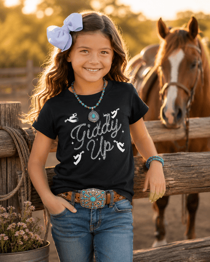 Young girl wearing a black t-shirt with 'Giddy Up' text, standing next to a horse in a rural setting.