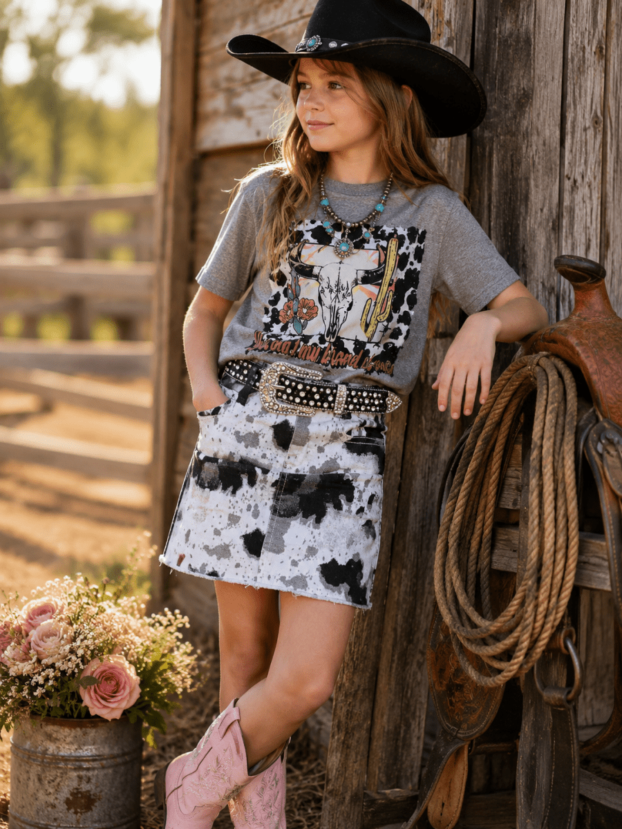 Young girl in cowboy attire standing next to a wooden wall with a floral arrangement and horse equipment.