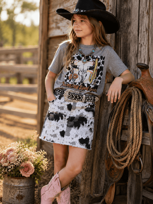 Young girl in cowboy attire standing next to a wooden wall with a floral arrangement and horse equipment.