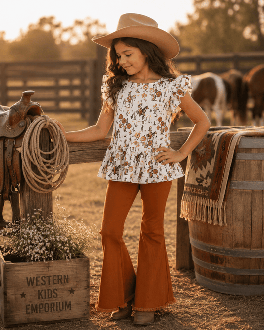 Young girl in a cowboy hat and flared pants standing in a rustic setting with a wooden barrel and horse equipment.