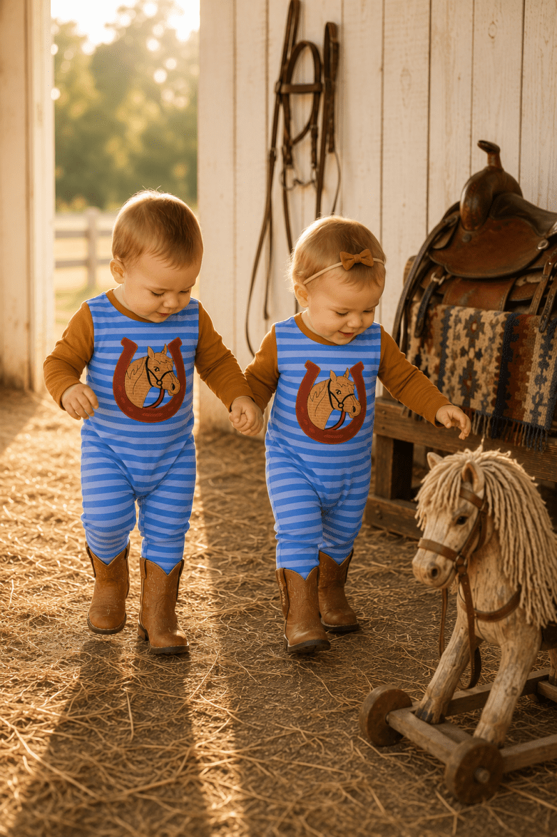 Two toddlers wearing matching blue and white striped one-piece outfits with a horse graphic on the chest, walking hand-in-hand in a barn with straw on the floor and a rocking horse nearby.