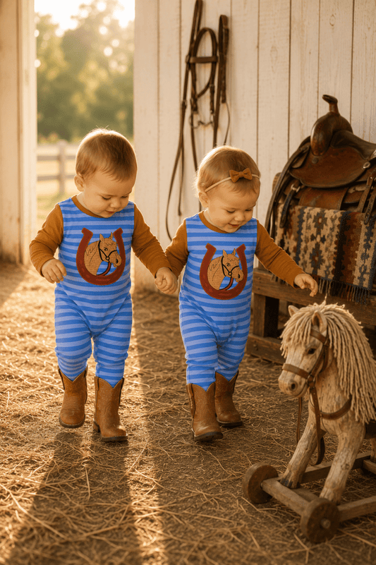 Two toddlers wearing matching blue and white striped one-piece outfits with a horse graphic on the chest, walking hand-in-hand in a barn with straw on the floor and a rocking horse nearby.