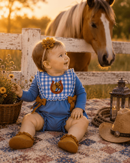 A baby wearing a blue long-sleeve romper with a blue-and-white gingham bib, sitting on a patterned blanket in a rustic outdoor setting with a horse behind a white fence.