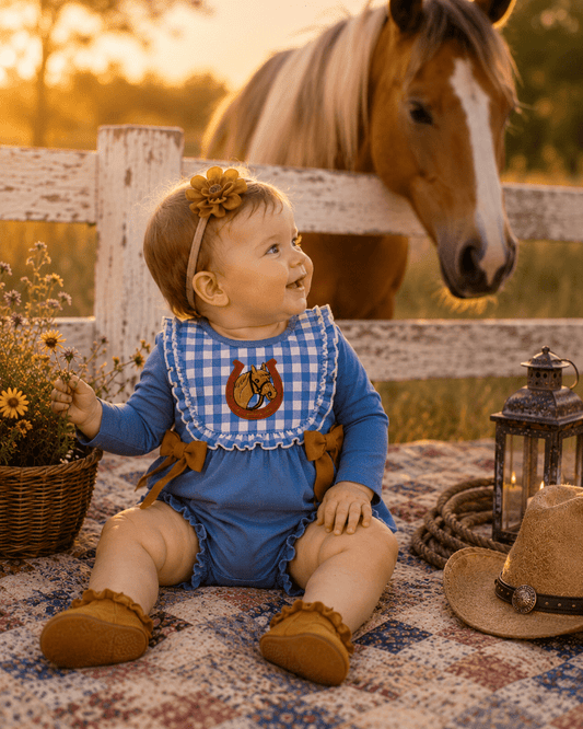 A baby wearing a blue long-sleeve romper with a blue-and-white gingham bib, sitting on a patterned blanket in a rustic outdoor setting with a horse behind a white fence.