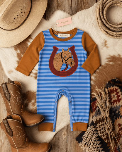 A baby romper laid flat on a cowhide rug with western-themed props including cowboy boots, a straw hat, rope, and a patterned scarf. The romper has blue and white horizontal stripes, tan sleeves and cuffs, and a large horseshoe-with-horse graphic on the chest.