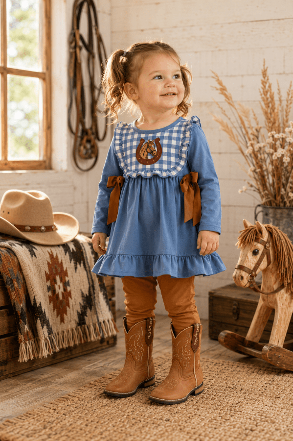 A young girl with pigtails stands in a rustic western-themed room wearing a blue long-sleeve dress with a gingham yoke, brown ribbon accents, tan leggings, and brown cowboy boots. A cowboy hat, blanket, rocking horse, and dried grass decor are visible around her.