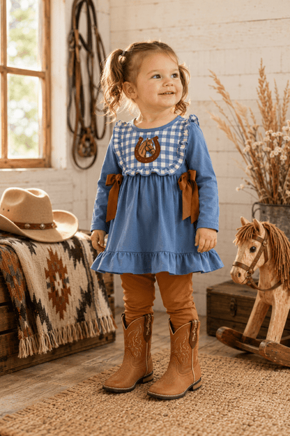 A young girl with pigtails stands in a rustic western-themed room wearing a blue long-sleeve dress with a gingham yoke, brown ribbon accents, tan leggings, and brown cowboy boots. A cowboy hat, blanket, rocking horse, and dried grass decor are visible around her.