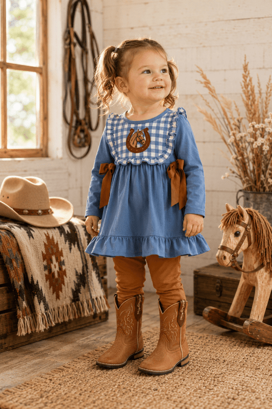 A young girl with pigtails stands in a rustic western-themed room wearing a blue long-sleeve dress with a gingham yoke, brown ribbon accents, tan leggings, and brown cowboy boots. A cowboy hat, blanket, rocking horse, and dried grass decor are visible around her.