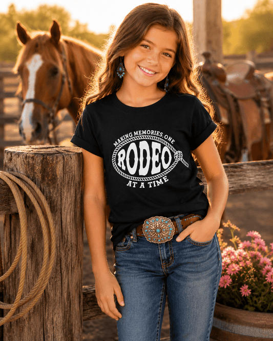 Young girl wearing a black t-shirt with 'Making Memories One Rodeo At A Time' text, standing in front of a horse and wooden fence.