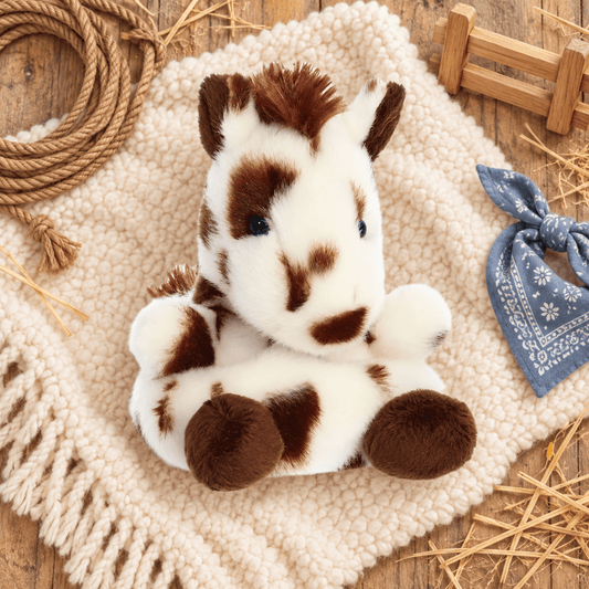 A plush toy horse with brown and white spots sits on a cream-colored textured blanket, surrounded by rustic props including a rope coil, wooden fence pieces, straw, and a blue bandana.