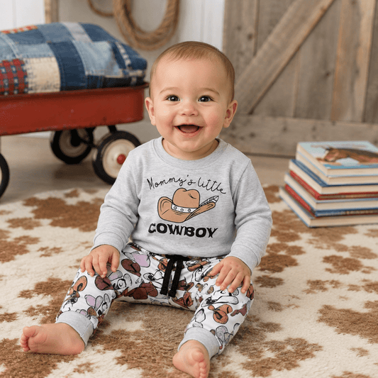 A smiling baby sitting on a patterned rug wearing a light gray long-sleeve shirt with a cowboy hat graphic and the words 'Mommy's little COWBOY,' paired with matching cowboy-print pants. A wagon and stacked books are visible in the room background.