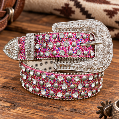 A western-style belt with a large engraved silver buckle, pink glitter base, and rows of clear rhinestones and silver studs, photographed on a rustic wooden surface.