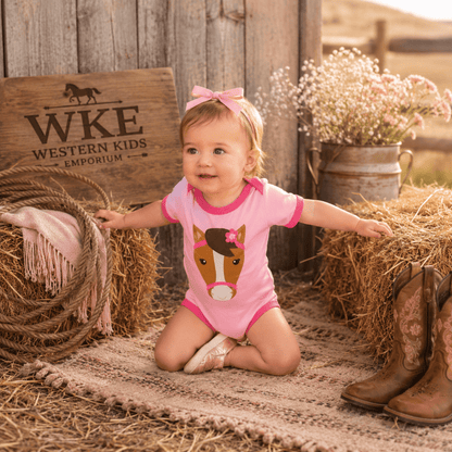 A toddler girl wearing a pink short-sleeve romper with a horse graphic sits on a straw rug in a rustic western-themed setting. She has a pink bow in her hair, with hay bales, cowboy boots, and a wooden sign reading 'WKE Western Kids Emporium' in the background.