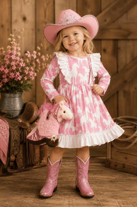 A young girl stands in a rustic barn-like setting wearing a pink and white patterned long-sleeve dress with ruffled trim, a matching pink cowboy hat, pink glittery cowboy boots, and carrying a pink stuffed horse and matching bag.