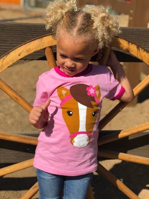 Child wearing a pink shirt with a horse design, standing in front of a wooden wheel.