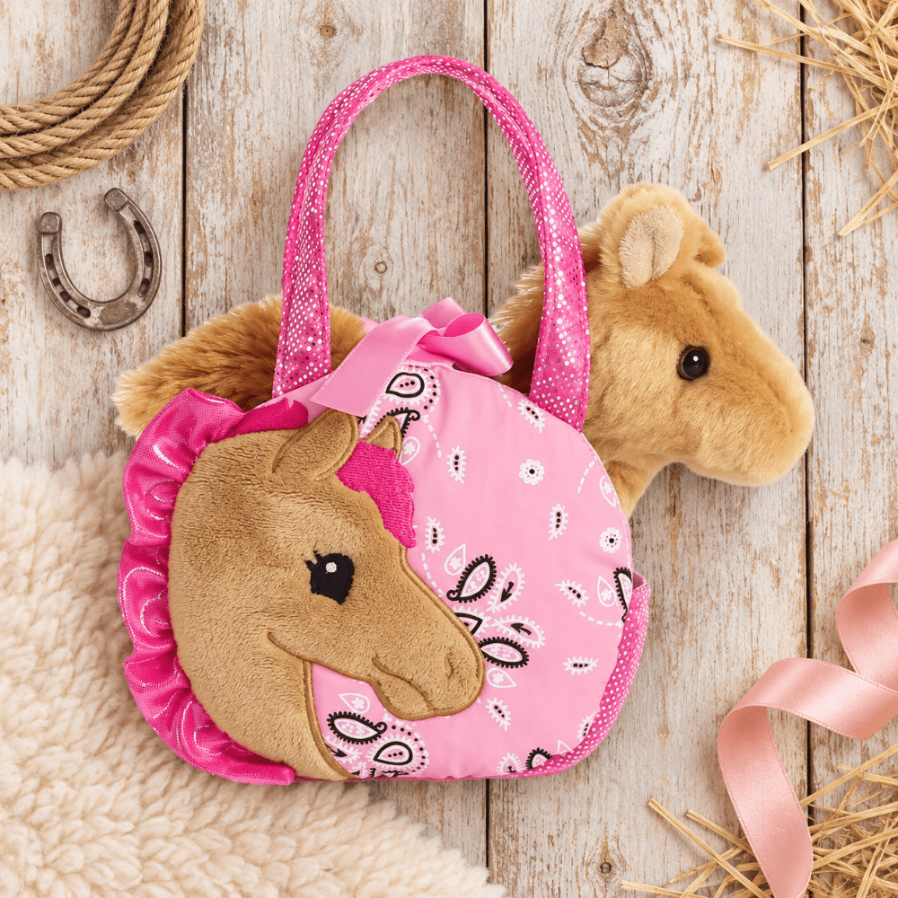 A pink plush horse-shaped handbag with a glittery pink handle, a brown plush horse face on the front, and a pink bandana bow, styled on a rustic white wood plank surface with rope, a horseshoe, and straw accents.