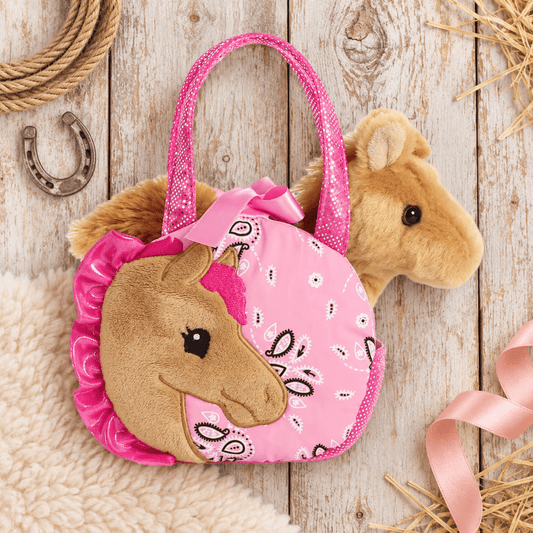 A pink plush horse-shaped handbag with a glittery pink handle, a brown plush horse face on the front, and a pink bandana bow, styled on a rustic white wood plank surface with rope, a horseshoe, and straw accents.
