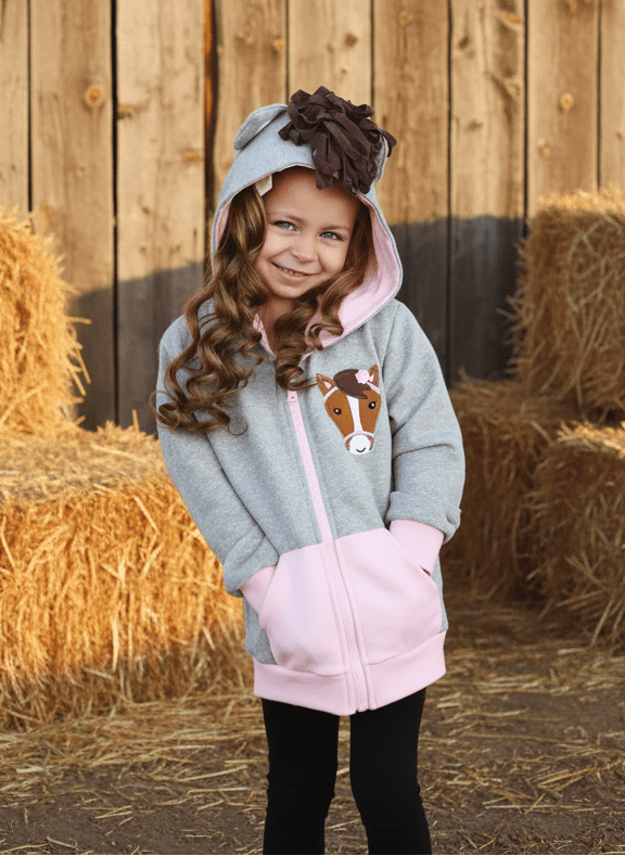 Child wearing a hoodie with a horse design in front of hay bales and a wooden wall.