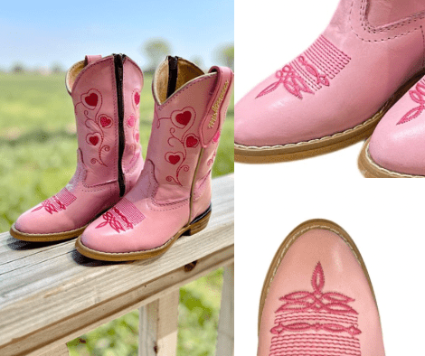 Pink children's cowboy boots with heart designs on a wooden surface and grass background.