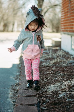 Child in a gray and pink horse hoodie and pants walking on a brick path outdoors.