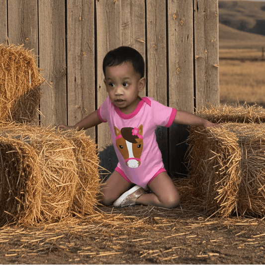 Child in a pink horse romper sitting on hay bales with a wooden fence and field in the background