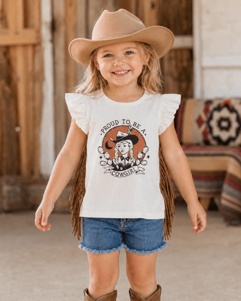 A young girl wearing a cream ruffle-sleeve graphic t-shirt with fringe, blue denim shorts, and a tan cowboy hat, standing in a rustic western-themed room.