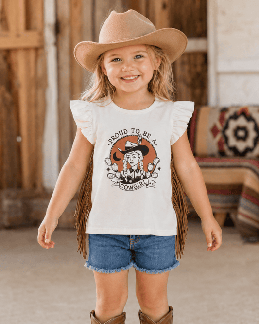 A young girl wearing a cream ruffle-sleeve graphic t-shirt with fringe, blue denim shorts, and a tan cowboy hat, standing in a rustic western-themed room.