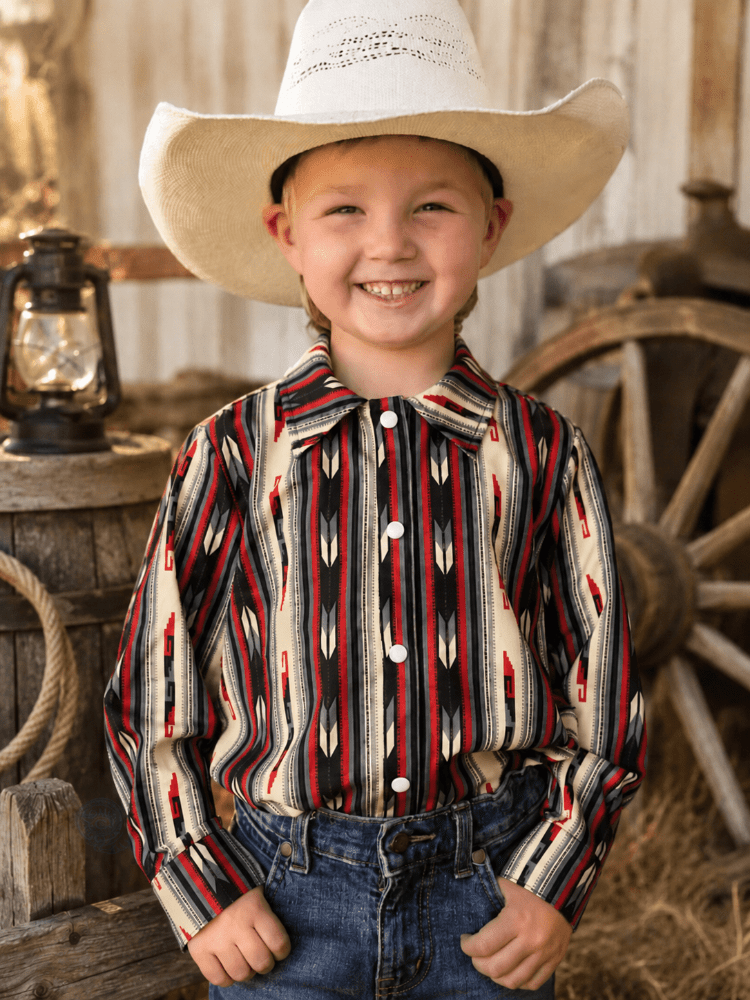 A young boy smiling and wearing a long-sleeve western shirt with a red, black, and cream geometric pattern, paired with a white cowboy hat and blue jeans, set against a rustic barn background.