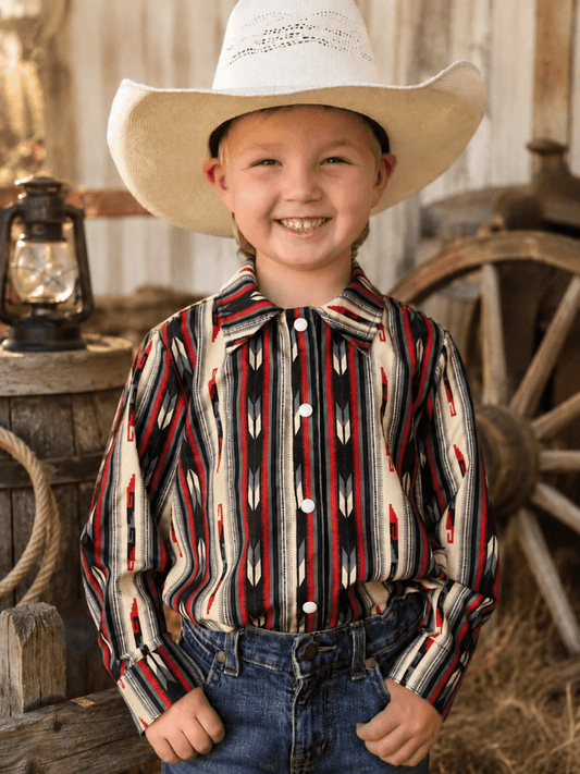 A young boy smiling and wearing a long-sleeve western shirt with a red, black, and cream geometric pattern, paired with a white cowboy hat and blue jeans, set against a rustic barn background.
