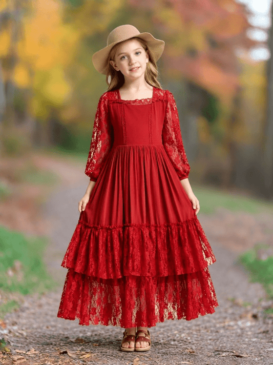 A young girl wearing a long red lace dress with three-quarter sleeves, a wide-brim tan hat, and brown sandals, standing on a dirt path with a blurred autumn forest background.