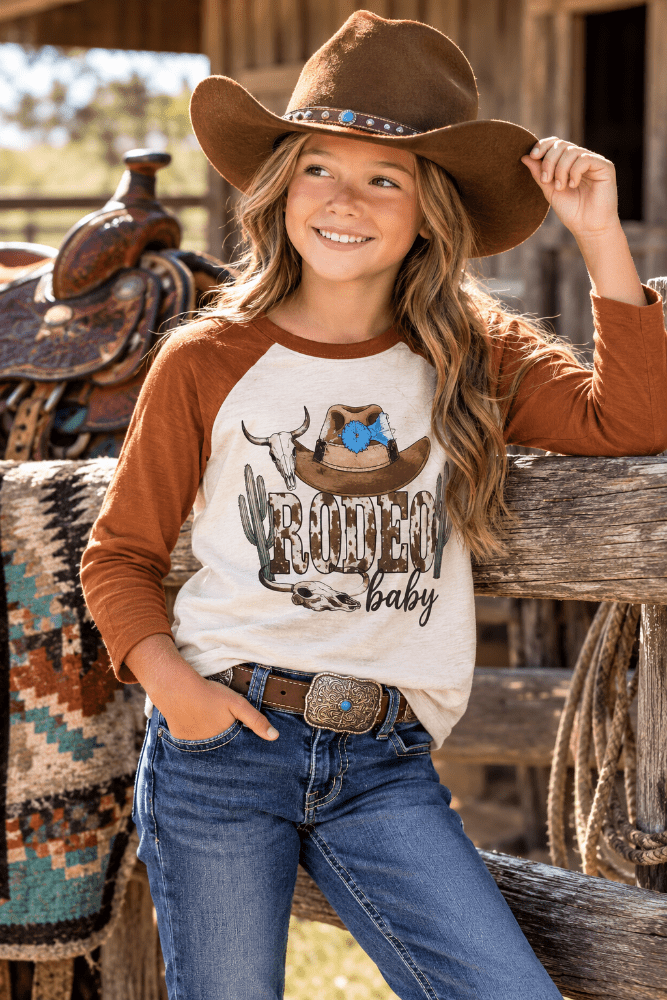 A young girl wearing a brown cowboy hat, a brown and cream raglan baseball t-shirt with a 'Roofer Baby' graphic, and blue jeans, posing on a rustic wooden fence in a western-themed setting.