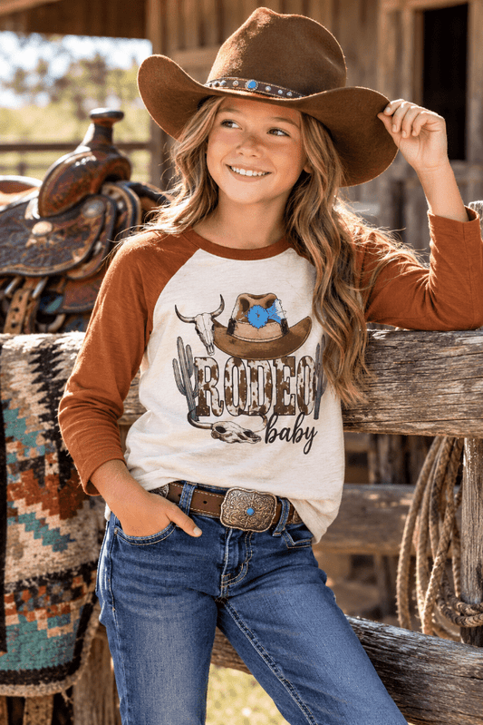 A young girl wearing a brown cowboy hat, a brown and cream raglan baseball t-shirt with a 'Roofer Baby' graphic, and blue jeans, posing on a rustic wooden fence in a western-themed setting.