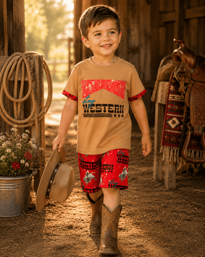 A young boy smiling in a rustic barn setting, wearing a tan short-sleeve t-shirt with a red and black 'Western' graphic, matching red patterned shorts, and brown cowboy boots while holding a hat.