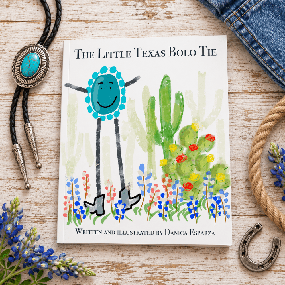 A children's book titled "The Little Texas Bolo Tie" lies on a whitewashed wooden surface surrounded by styled props including a turquoise bolo tie, blue flowers, rope, a horseshoe, and denim. The cover shows a smiling blue flower-like figure standing among cacti and blue/red flowers.