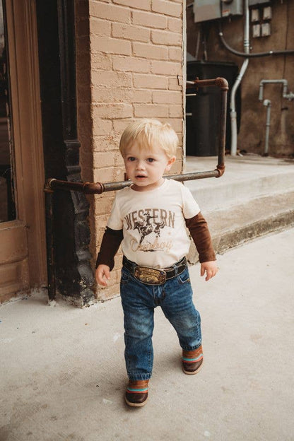 A toddler walking outdoors in front of a brick building, wearing a cream western-style graphic t-shirt with brown sleeves, blue jeans, a brown belt, and striped boots.
