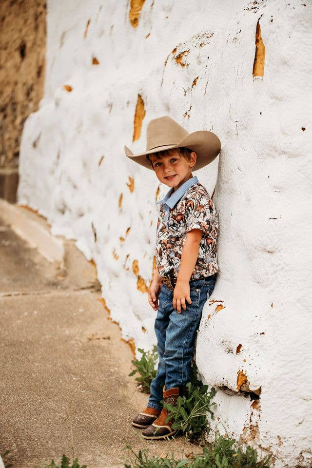 Child wearing a cowboy hat and patterned shirt leaning against a textured white wall.