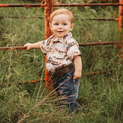 Child standing in a grassy field, wearing his train cowboy shirt next to a rusted fence