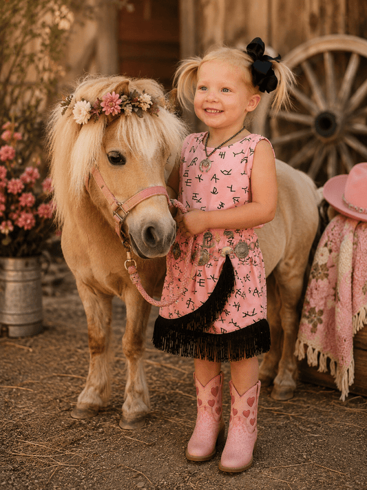 Young girl in a pink dress standing next to a small horse with a floral wreath, in front of a wooden wagon wheel.