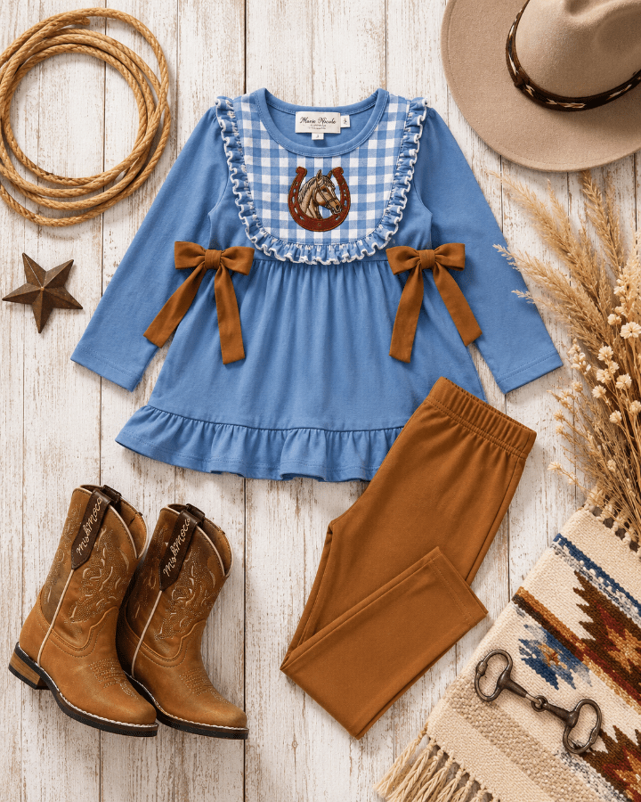 A children's western-style outfit set laid flat on a whitewashed wood surface, including a light blue long-sleeve top with a gingham yoke, brown bow details, a horse motif at the neckline, matching brown pants, tan cowboy boots, a straw hat, rope, and dried floral decor.