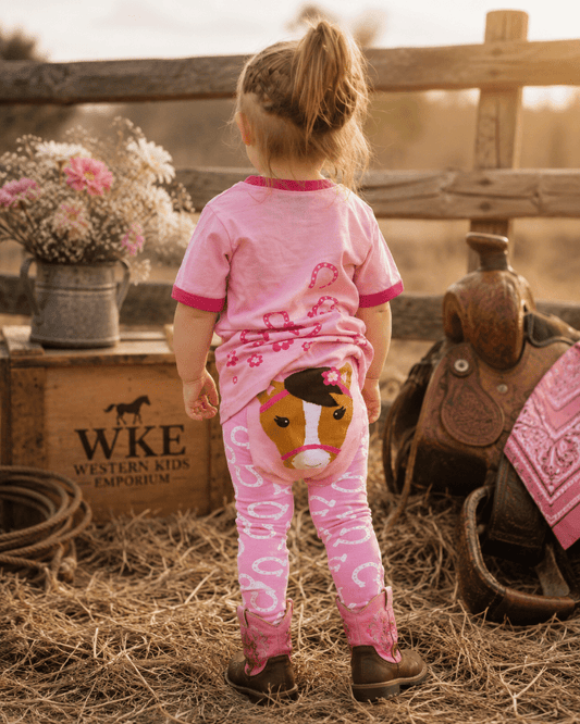 A toddler stands with their back to the camera in a rustic western-themed setting, wearing a pink short-sleeve t-shirt and matching pink leggings with white patterns and a cartoon horse graphic on the back.