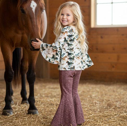 A young girl with long blonde hair stands in a barn next to a brown horse, wearing a white long-sleeve top with a green and brown floral pattern and dusty rose bell-bottom pants.