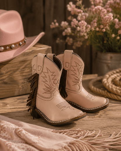 A pair of light tan western-style leather cowboy boots with fringe and embroidered stitching, displayed on a rustic wooden surface with a matching pink hat, flowers, and rope in the background.
