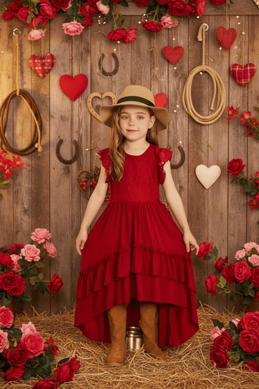 Young girl in a red dress and straw hat standing in front of a wooden wall with floral decorations.
