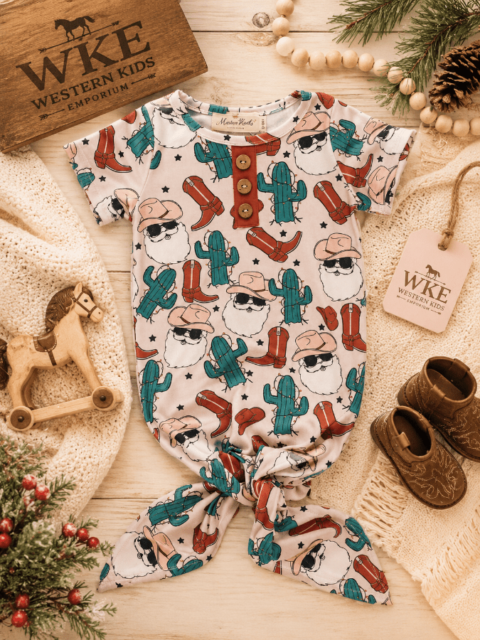 A baby romper and matching bootie set with a Western Santa Claus cactus print, styled in a flat lay on a light wood background with cozy props including a wooden horse, knit blanket, and WKE branding.