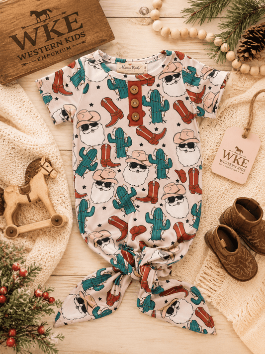 A baby romper and matching bootie set with a Western Santa Claus cactus print, styled in a flat lay on a light wood background with cozy props including a wooden horse, knit blanket, and WKE branding.